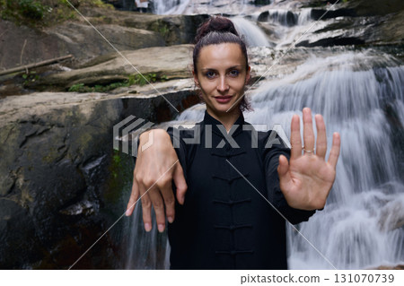 Woman practicing Tai Chi in nature, wearing a traditional black martial arts uniform, standing gracefully near a waterfall, focused on meditation and balance. Healthy lifestyle in asian culture 131070739