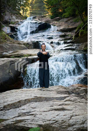 Beautiful young woman performing slow tai chi movements near a waterfall and river at sunset capturing harmony balance and fitness in nature. 131070749
