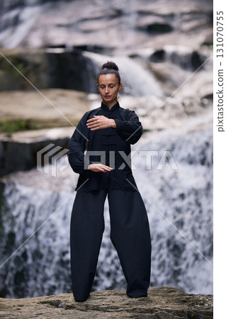 Woman practicing Tai Chi in nature, wearing a traditional black martial arts uniform, standing gracefully near a waterfall, focused on meditation and balance. Healthy lifestyle in asian culture Woman practicing Tai Chi in nature, wearing a traditional black martial arts uniform, standing gracefully near a waterfall, focused on meditation and balance. Healthy lifestyle in asian culture 131070755