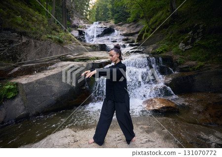 Woman practicing Tai Chi in nature, wearing a traditional black martial arts uniform, standing gracefully near a waterfall, focused on meditation and balance. Healthy lifestyle in asian culture Woman practicing Tai Chi in nature, wearing a traditional black martial arts uniform, standing gracefully near a waterfall, focused on meditation and balance. Healthy lifestyle in asian culture 131070772