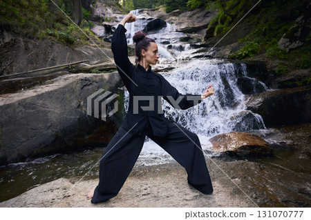 Woman practicing Tai Chi in nature, wearing a traditional black martial arts uniform, standing gracefully near a waterfall, focused on meditation and balance. Healthy lifestyle in asian culture 131070777