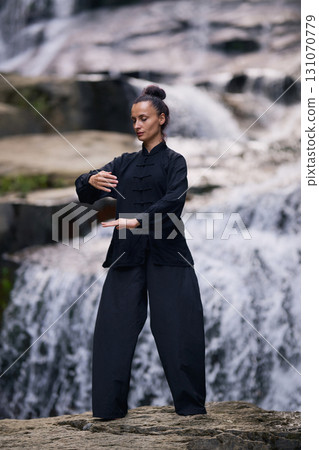 Woman practicing Tai Chi in nature, wearing a traditional black martial arts uniform, standing gracefully near a waterfall, focused on meditation and balance. Healthy lifestyle in asian culture Woman practicing Tai Chi in nature, wearing a traditional black martial arts uniform, standing gracefully near a waterfall, focused on meditation and balance. Healthy lifestyle in asian culture 131070779