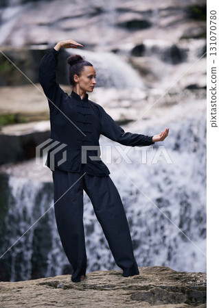Woman practicing Tai Chi in nature, wearing a traditional black martial arts uniform, standing gracefully near a waterfall, focused on meditation and balance. Healthy lifestyle in asian culture 131070780