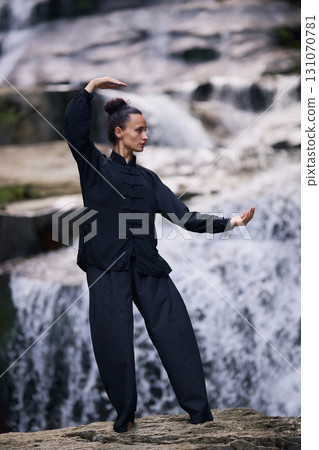 Woman practicing Tai Chi in nature, wearing a traditional black martial arts uniform, standing gracefully near a waterfall, focused on meditation and balance. Healthy lifestyle in asian culture Woman practicing Tai Chi in nature, wearing a traditional black martial arts uniform, standing gracefully near a waterfall, focused on meditation and balance. Healthy lifestyle in asian culture 131070781