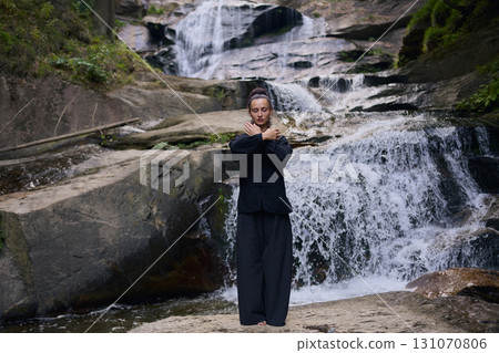 Woman practicing Tai Chi in nature, wearing a traditional black martial arts uniform, standing gracefully near a waterfall, focused on meditation and balance. Healthy lifestyle in asian culture 131070806