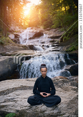 A woman sits in lotus pose, practicing yoga and tai chi under a tropical waterfall, meditating with calm strength and mindfulness, fully connected to the energy of nature, tranquility and inner 131070807