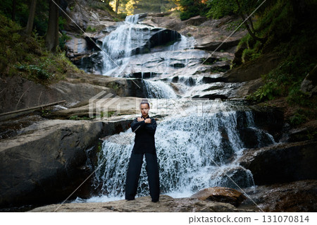 Beautiful young woman performing slow tai chi movements near a waterfall and river at sunset capturing harmony balance and fitness in nature. 131070814