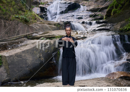 Woman practicing Tai Chi in nature, wearing a traditional black martial arts uniform, standing gracefully near a waterfall, focused on meditation and balance. Healthy lifestyle in asian culture 131070836