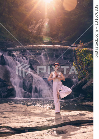 Woman doing yoga tree pose in front of waterfall at sunrise peaceful meditation in nature Woman doing yoga tree pose in front of waterfall at sunrise peaceful meditation in nature 131070838
