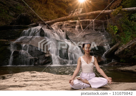 Young woman sitting in lotus pose under tropical waterfall meditating with calm strength and mindfulness fully connected to nature energy tranquility and inner balance Young woman sitting in lotus pose under tropical waterfall meditating with calm strength and mindfulness fully connected to nature energy tranquility and inner balance 131070842
