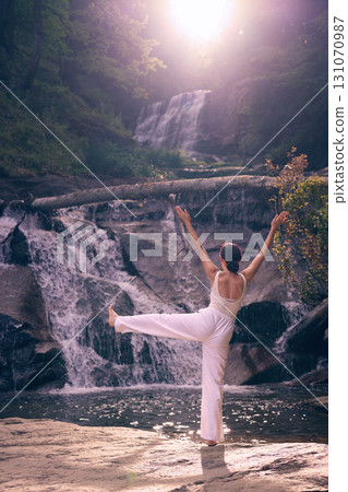 Woman doing yoga tree pose in front of waterfall at sunrise peaceful meditation in nature 131070987