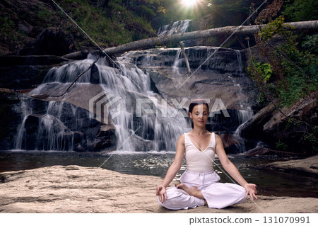 Young woman sitting in lotus pose under tropical waterfall meditating with calm strength and mindfulness fully connected to nature energy tranquility and inner balance 131070991