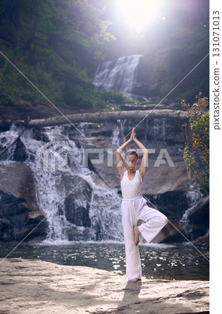 Woman doing yoga tree pose in front of waterfall at sunrise peaceful meditation in nature Woman doing yoga tree pose in front of waterfall at sunrise peaceful meditation in nature 131071013