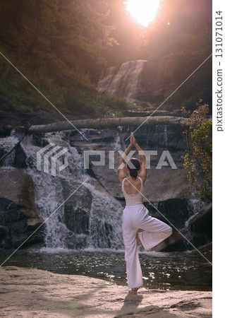 Woman doing yoga tree pose in front of waterfall at sunrise peaceful meditation in nature Woman doing yoga tree pose in front of waterfall at sunrise peaceful meditation in nature 131071014