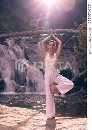 Woman doing yoga tree pose in front of waterfall at sunrise peaceful meditation in nature Woman doing yoga tree pose in front of waterfall at sunrise peaceful meditation in nature 131071067