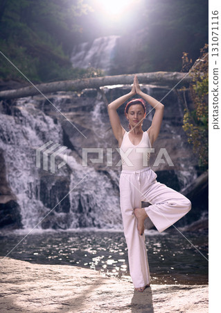 Woman doing yoga tree pose in front of waterfall at sunrise peaceful meditation in nature 131071116