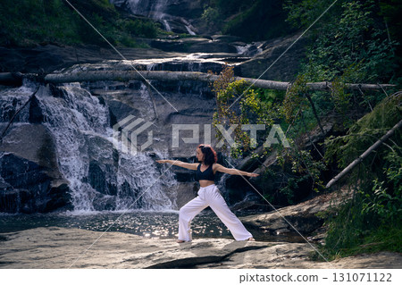 Woman practicing yoga warrior pose near waterfall at sunrise peaceful meditation in nature outdoor fitness Woman practicing yoga warrior pose near waterfall at sunrise peaceful meditation in nature outdoor fitness 131071122