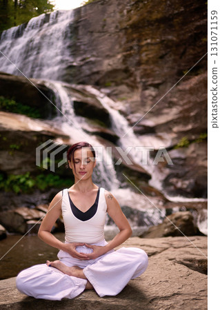 Young woman sitting in lotus pose under tropical waterfall meditating with calm strength and mindfulness fully connected to nature energy tranquility and inner balance 131071159
