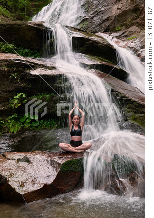 Young woman sitting in lotus pose under tropical waterfall meditating with calm strength and mindfulness fully connected to nature energy tranquility and inner balance 131071187