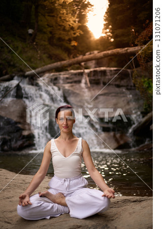Young woman sitting in lotus pose under tropical waterfall meditating with calm strength and mindfulness fully connected to nature energy tranquility and inner balance 131071206