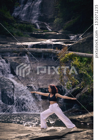 Woman practicing yoga warrior pose near waterfall at sunrise peaceful meditation in nature outdoor fitness 131071235
