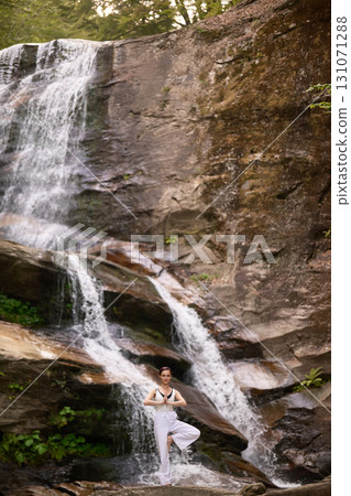 Woman doing yoga tree pose in front of waterfall at sunrise peaceful meditation in nature 131071288