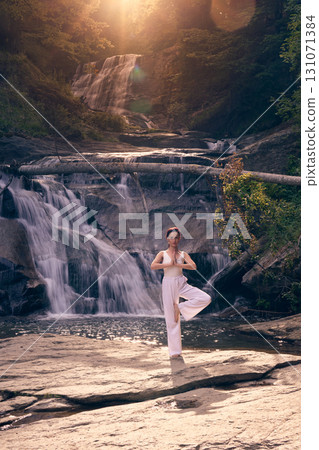 Woman doing yoga tree pose in front of waterfall at sunrise peaceful meditation in nature Woman doing yoga tree pose in front of waterfall at sunrise peaceful meditation in nature 131071384