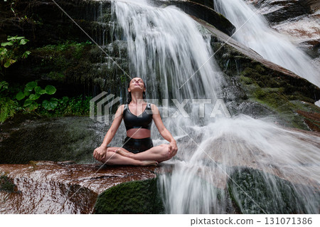 Young woman sitting in lotus pose under tropical waterfall meditating with calm strength and mindfulness fully connected to nature energy tranquility and inner balance Young woman sitting in lotus pose under tropical waterfall meditating with calm strength and mindfulness fully connected to nature energy tranquility and inner balance 131071386