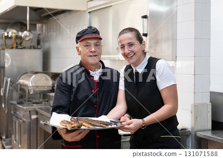 Two chefs in black uniforms working together while serving freshly fried fish in a commercial kitchen. Concept of seafood preparation, teamwork, culinary skills, and hospitality industry. 131071588