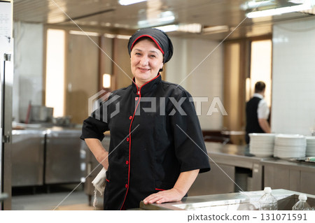 Portrait of a female chef in a black uniform with red details smiling in a professional restaurant kitchen. Image highlights hospitality staff, confidence, and culinary professionalism. Portrait of a female chef in a black uniform with red details smiling in a professional restaurant kitchen. Image highlights hospitality staff, confidence, and culinary professionalism. 131071591