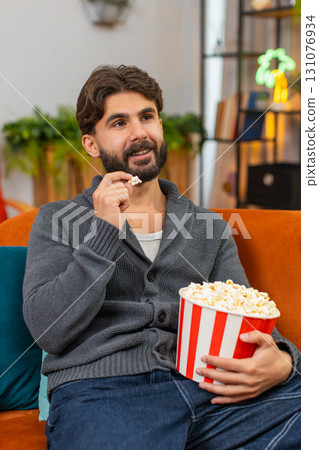 Indian man at home sofa watches series finale on TV holds popcorn eating, excited and smiles Indian man at home sofa watches series finale on TV holds popcorn eating, excited and smiles 131076934