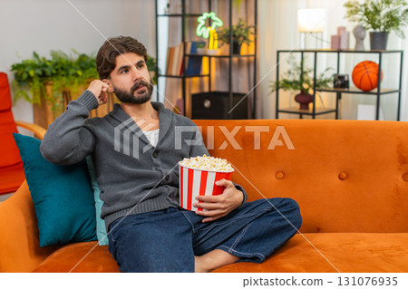 Indian man at home sofa watches series finale on TV holds popcorn eating, excited and smiles Indian man at home sofa watches series finale on TV holds popcorn eating, excited and smiles 131076935