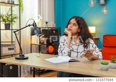 Indian woman checks smartphone info cross referencing notebook studying data research indoors focus Indian woman checks smartphone info cross referencing notebook studying data research indoors focus 131077084