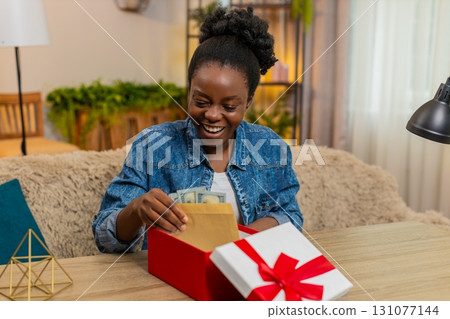 African American young woman opens birthday gift box with envelope of cash dollars sitting at home African American young woman opens birthday gift box with envelope of cash dollars sitting at home 131077144