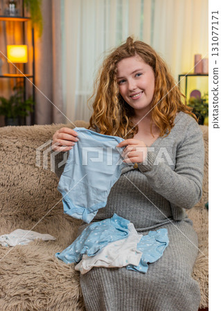 Pregnant woman sorting baby clothes at home, smiling emotional expression, preparing for motherhood 131077171