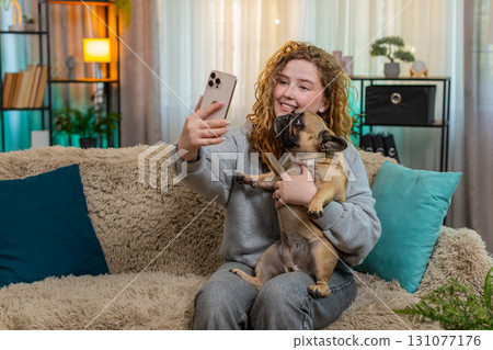 Young woman taking selfie with pug dog on home sofa holding smartphone and smiling joyfully with pet 131077176