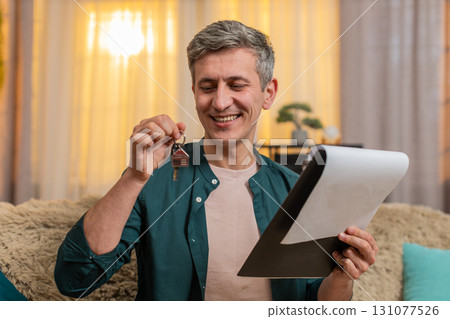 Young man smiles proudly holding keys documents after signing contract for new apartment at home 131077526