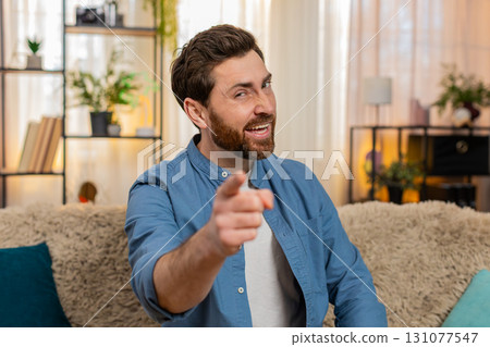 Young man playfully winking flirting smiling pointing at camera cheerful happy charming home sofa Young man playfully winking flirting smiling pointing at camera cheerful happy charming home sofa 131077547
