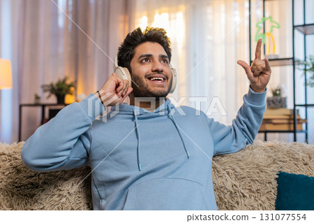 Indian young man sitting on sofa listening to music with headphones dancing to rhythm at home Indian young man sitting on sofa listening to music with headphones dancing to rhythm at home 131077554