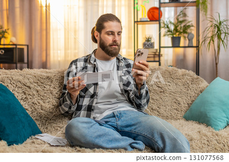 Young man sorting bills at home, sofa budget check, phone in hand, multiple papers, calm focus vibe Young man sorting bills at home, sofa budget check, phone in hand, multiple papers, calm focus vibe 131077568