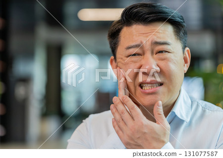 Chinese businessman in office lobby working on laptop and feeling toothache in modern office lobby Chinese businessman in office lobby working on laptop and feeling toothache in modern office lobby 131077587
