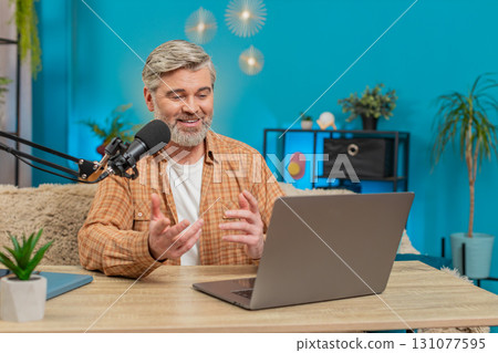 Mature man at home table speaking into microphone during live online stream, responding with energy 131077595