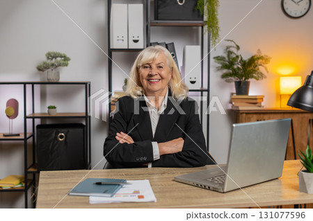Smiling senior businesswoman using laptop computer and looking at camera at home office table Smiling senior businesswoman using laptop computer and looking at camera at home office table 131077596