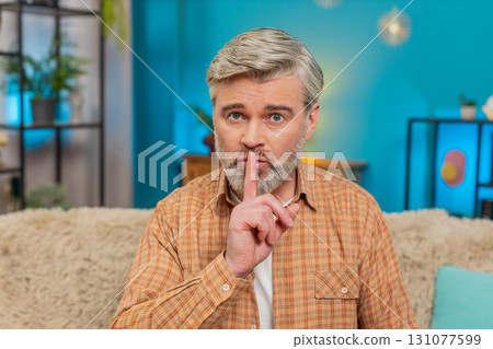 Mature man sitting on sofa making quiet gesture, expressing seriousness and secrecy at home Mature man sitting on sofa making quiet gesture, expressing seriousness and secrecy at home 131077599