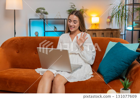 Happy young woman making online video call on laptop talking waving hand hello hi sitting on sofa Happy young woman making online video call on laptop talking waving hand hello hi sitting on sofa 131077609