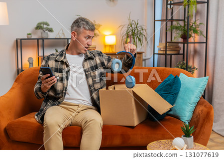 Unhappy man unpacking carton box feeling dissatisfied with getting damaged wireless headphones home 131077619