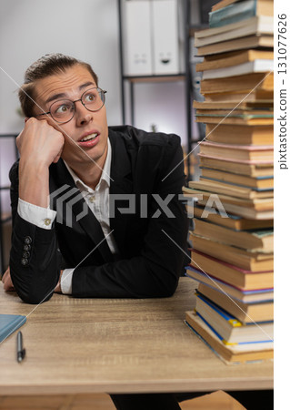 Tired exhausted stressed businessman looking at large pile of books sitting at home office desk 131077626