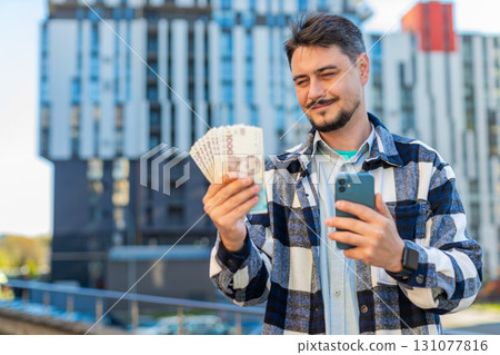 Happy rich mature business man using smartphone and counting money hryvnias on downtown city street 131077816