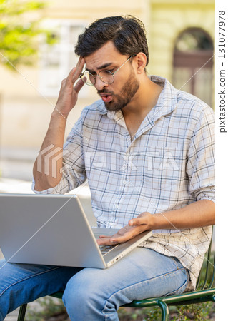 Displeased Indian man using laptop working, loses becoming surprised sudden lottery results outdoors Displeased Indian man using laptop working, loses becoming surprised sudden lottery results outdoors 131077978
