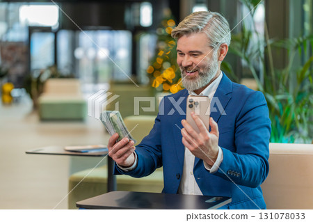 Successful businessman holding smartphone and counting dollar bill money fan sitting in office lobby Successful businessman holding smartphone and counting dollar bill money fan sitting in office lobby 131078033